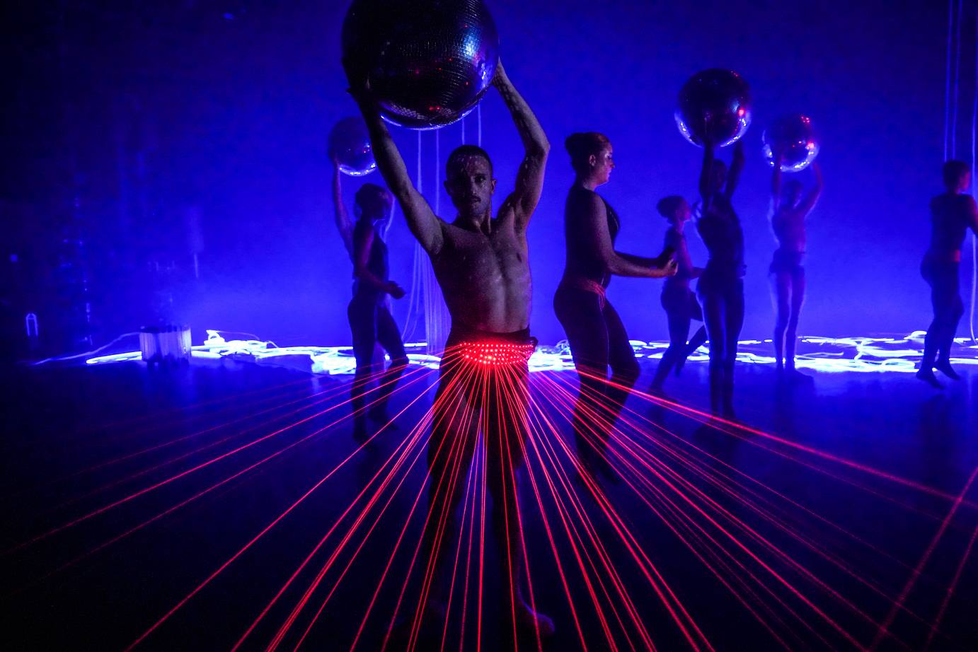 a chorus of dancers in a blue lit room, white neon lights lay on the floor in the background,some dancers hold large disco balls others dont, a central figure stares out at us holding a huge disco ball with red lights emitted from his waist band
